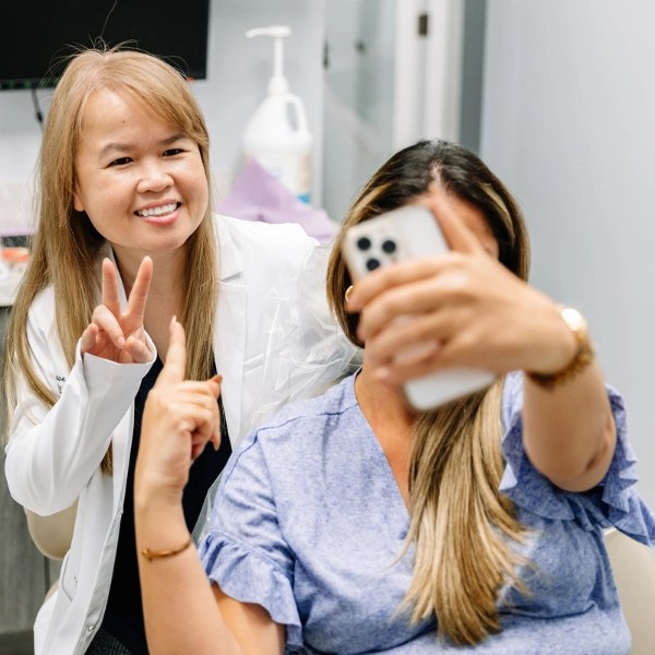 Dr. Phuong-Uyen Le in a white lab coat makes a peace sign while a patient in a purple shirt takes a selfie with her on a smartphone.